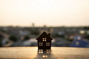Silhouette of a small house model on a ledge, with a soft-focus townscape and warm, golden sunset in the background, conveying a peaceful, dreamy mood.